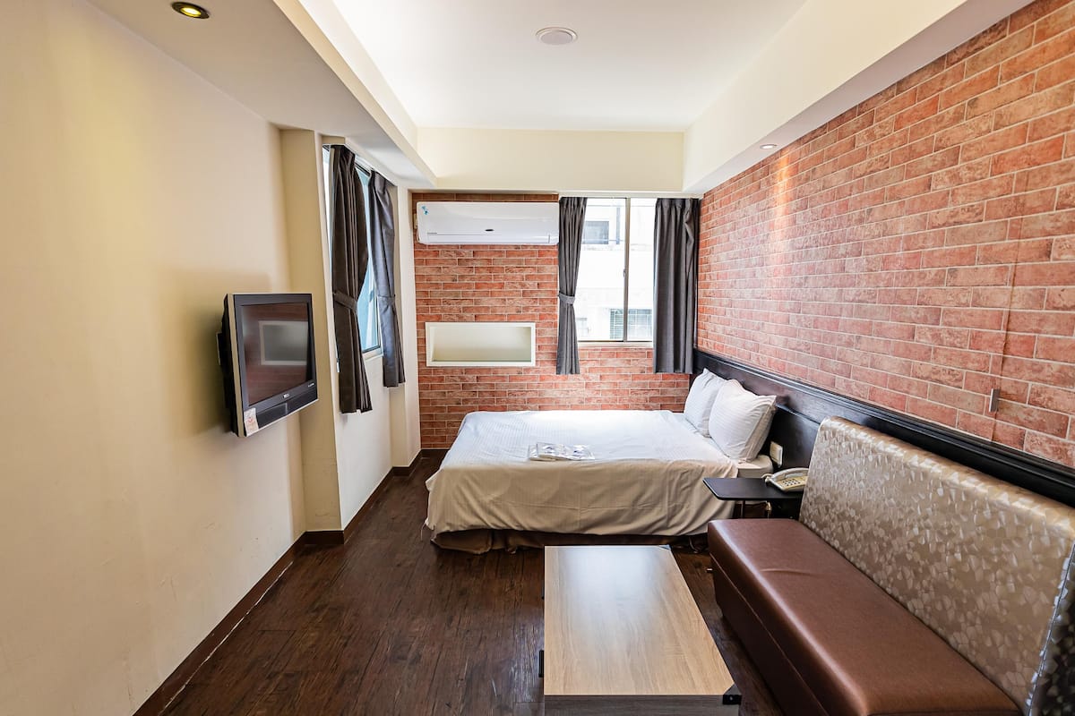 Ein kleines Hotelzimmer mit Bett, Sitzbank, Fernseher und einer Ziegelwand.