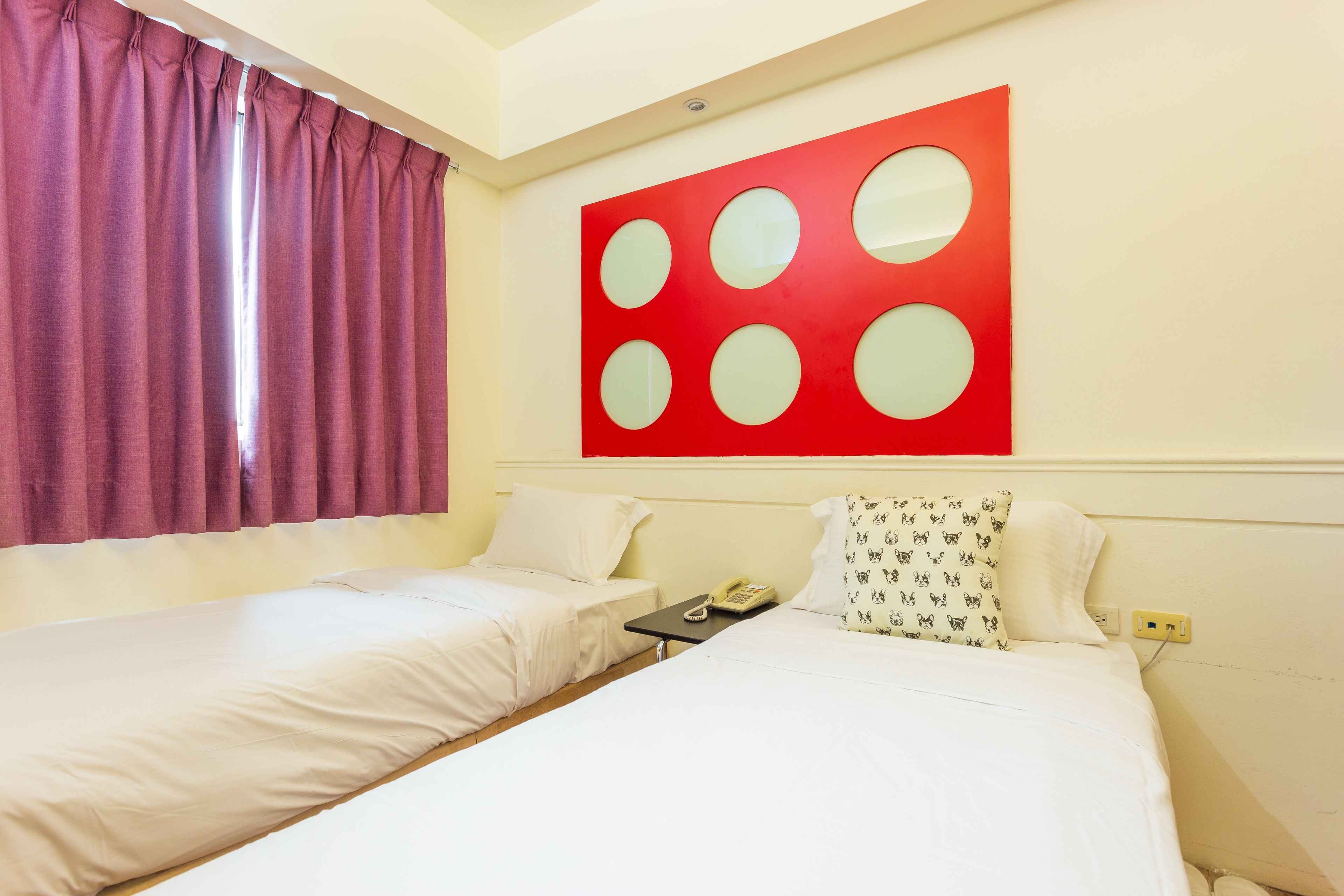 Habitación de hotel con dos camas individuales, una obra de arte roja en la pared, un teléfono en la mesita de noche y una ventana con cortinas moradas.