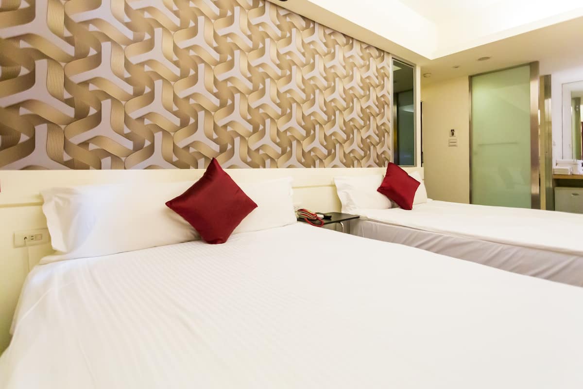 Ein Hotelzimmer mit zwei Betten, jeweils mit weißen Laken und roten Kissen. Das Zimmer zeichnet sich durch ein modernes Wanddesign und eine sichtbare Badezimmertür aus.