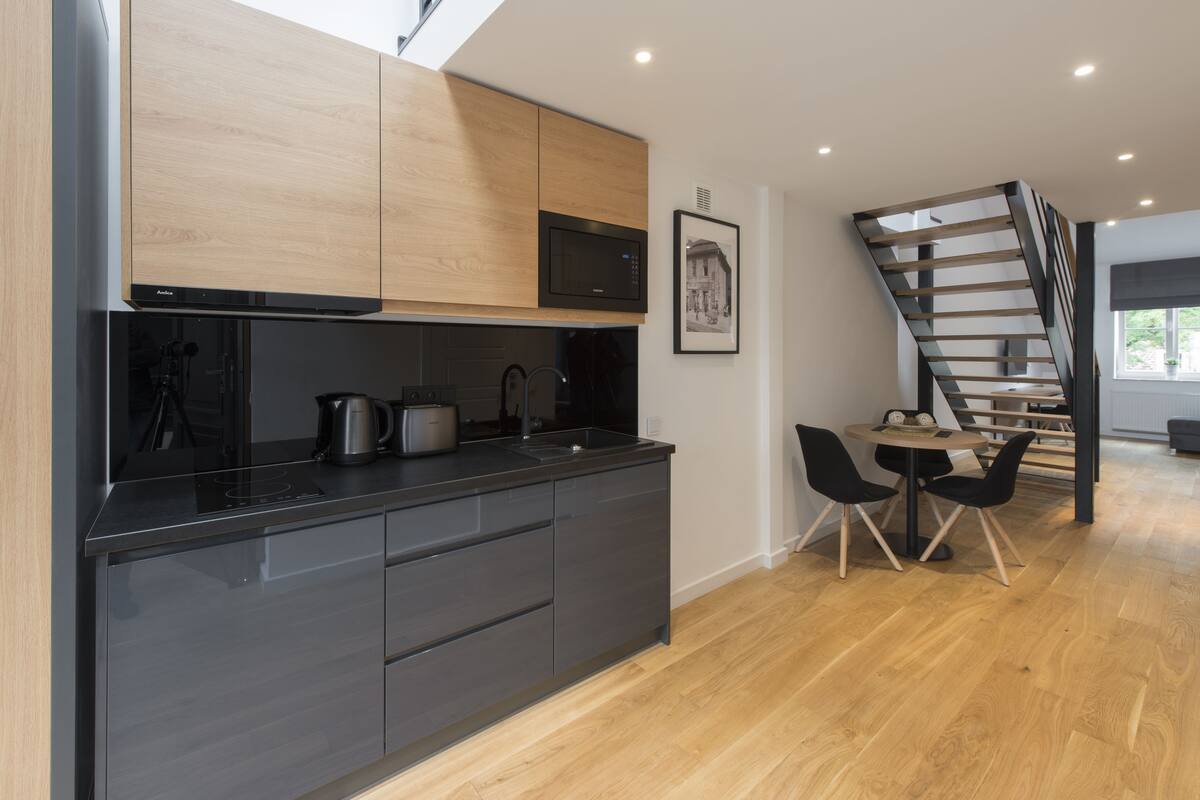 A modern kitchen with dark cabinetry, a microwave, and a kettle on the counter. There is a dining area with a table and chairs, and a staircase leading to another level.