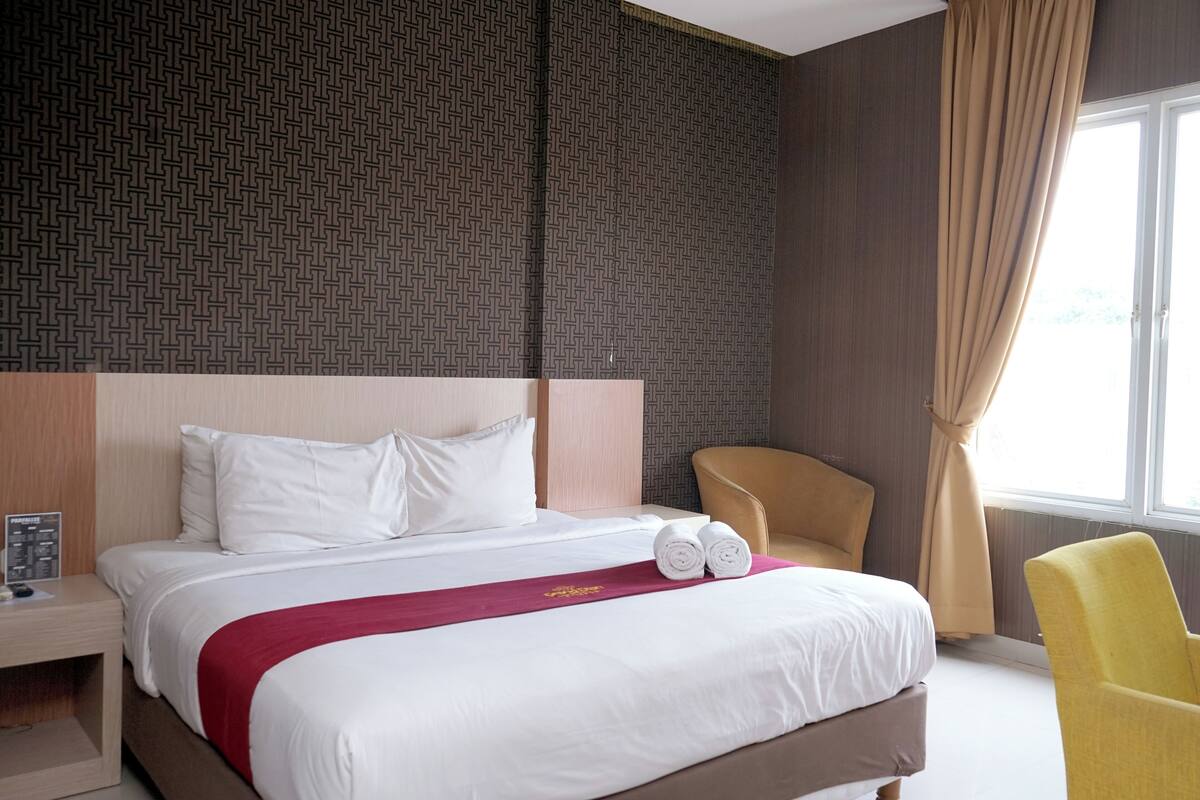 A hotel room with a large bed, a wooden headboard, a red throw blanket, and a yellow armchair.