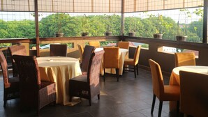 A restaurant with wicker chairs and tables, large windows with a view of greenery, and potted plants on the windowsill. - Grand Dian Boutique Hotel (North Cirebon)