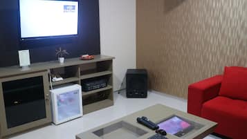 A living room with a flat-screen TV, a small refrigerator, a red sofa, and a coffee table with electronic devices.