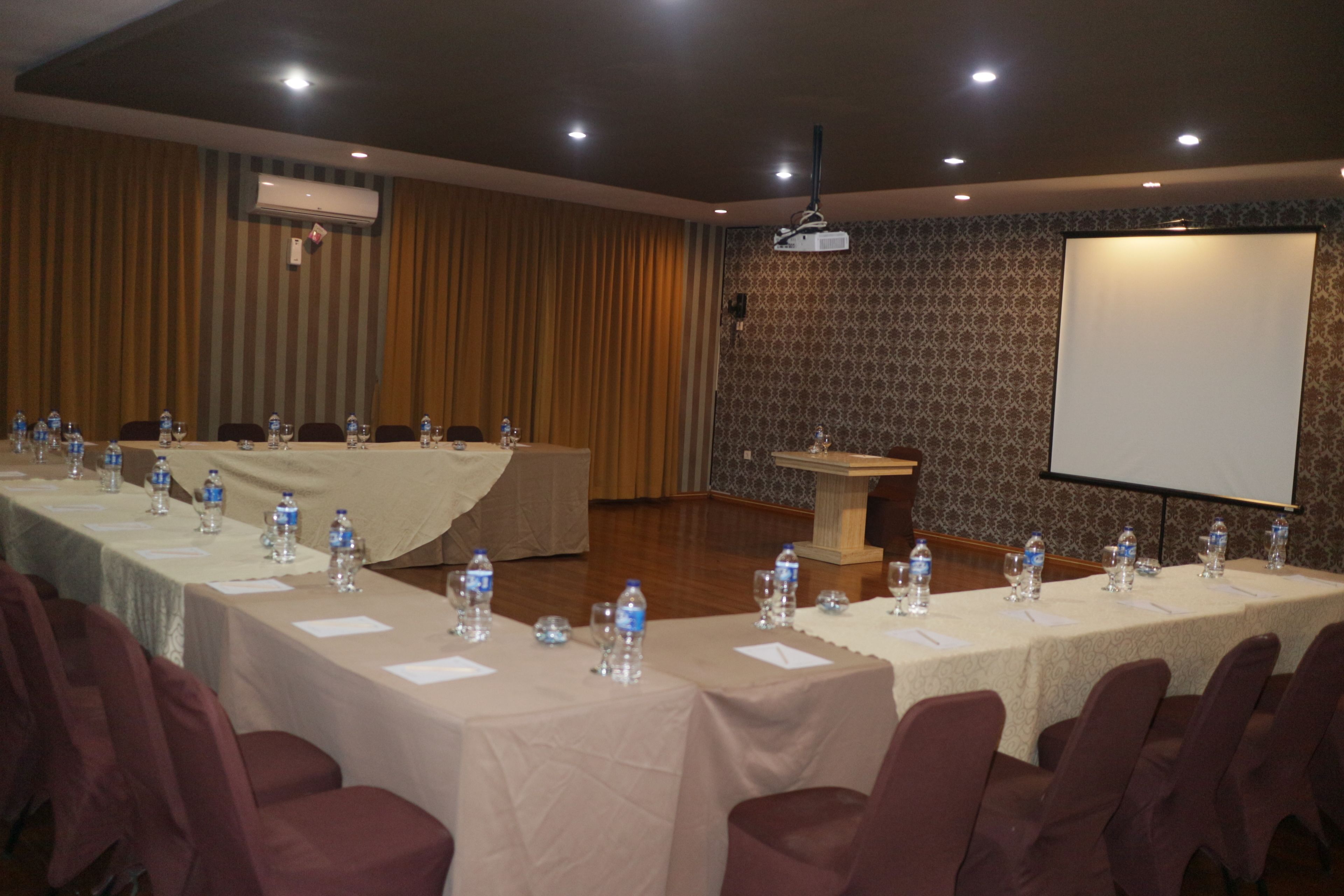 Un salón de conferencias con mesas largas, sillas, botellas de agua y una pantalla de proyector.