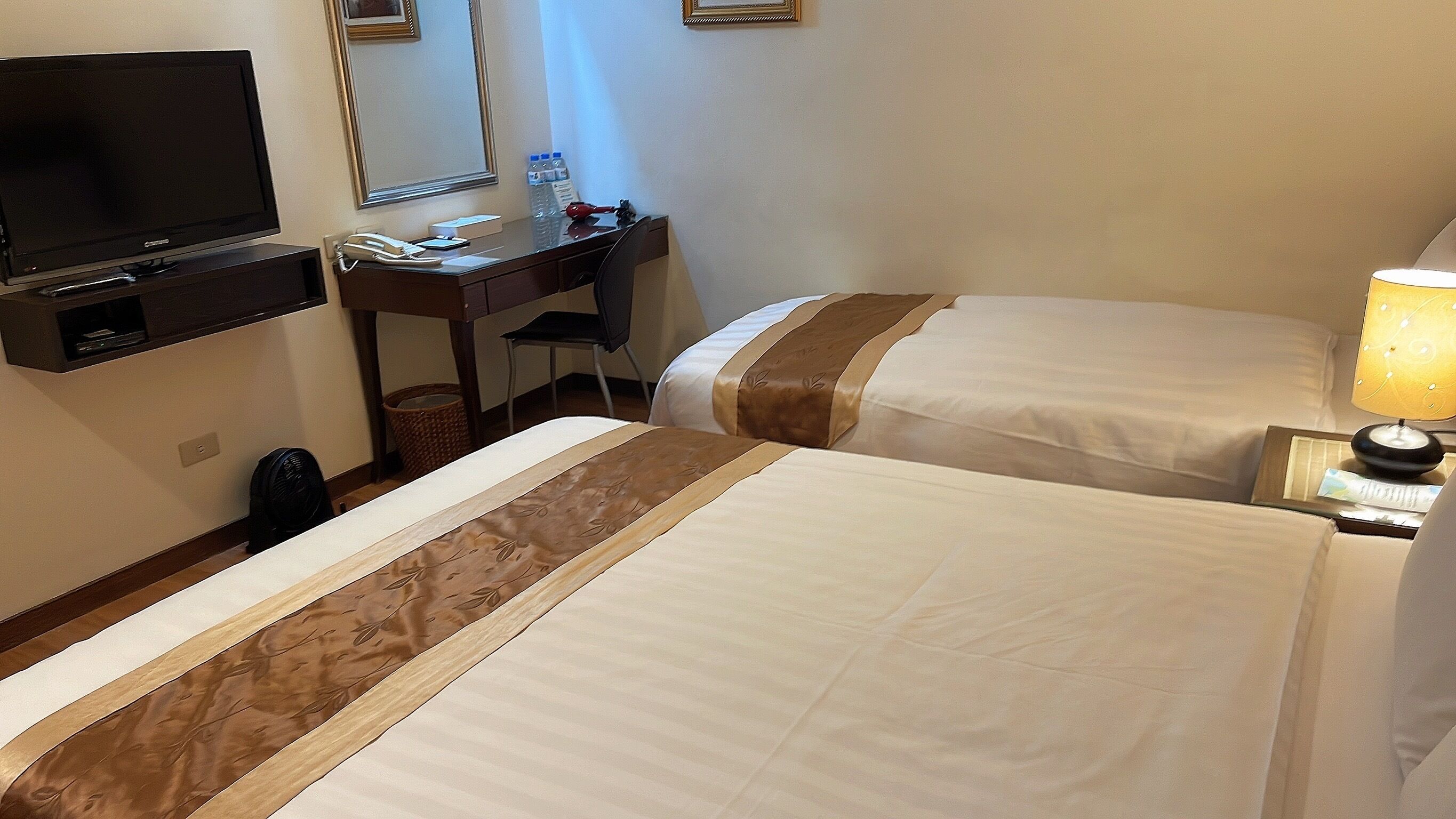 Quarto de hotel com duas camas, uma escrivaninha com computador e telefone, uma televisão e um abajur.