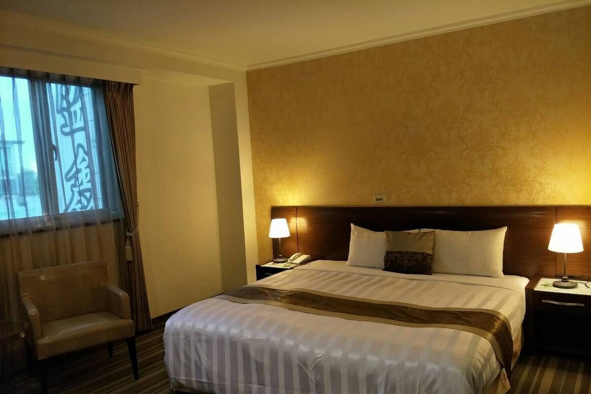 A hotel room with a large bed, two bedside tables with lamps, a chair, and a window with curtains.