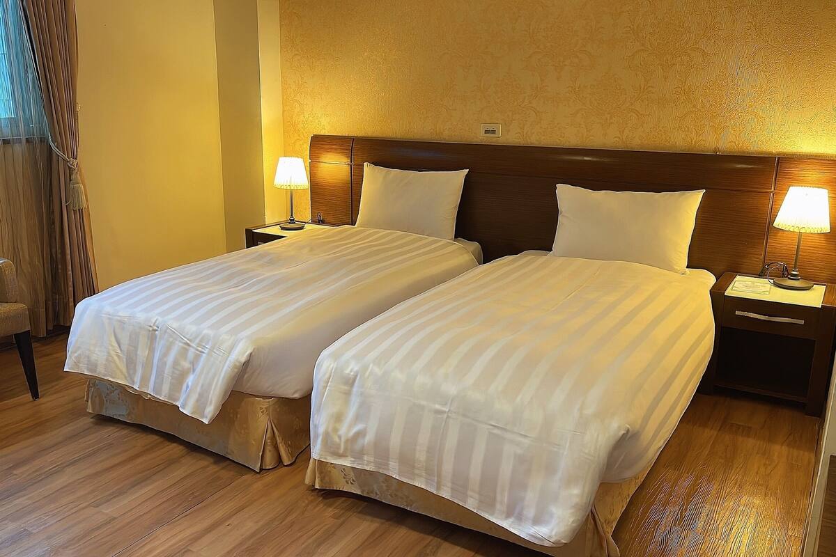 A hotel room with two beds, wooden flooring, a chair, and bedside lamps.