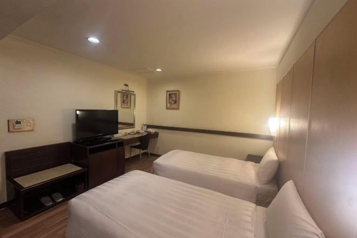A hotel room with two beds, a desk with a chair, a television, and a small shelf.
