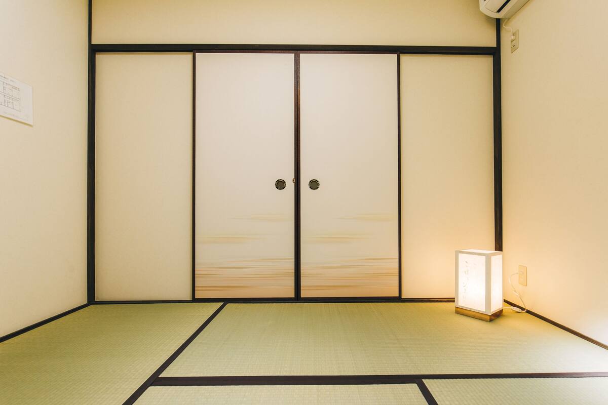 Una habitación japonesa tradicional con suelo de tatami, puertas correderas y una mesita de noche con lámpara.