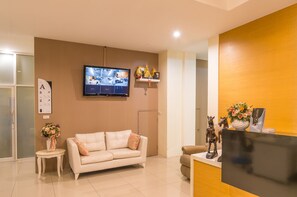 A modern reception area with a sofa, a TV displaying a live feed, a small table with a vase, and a reception desk with a flower arrangement. - Alisa Krabi Hotel (Krabi)