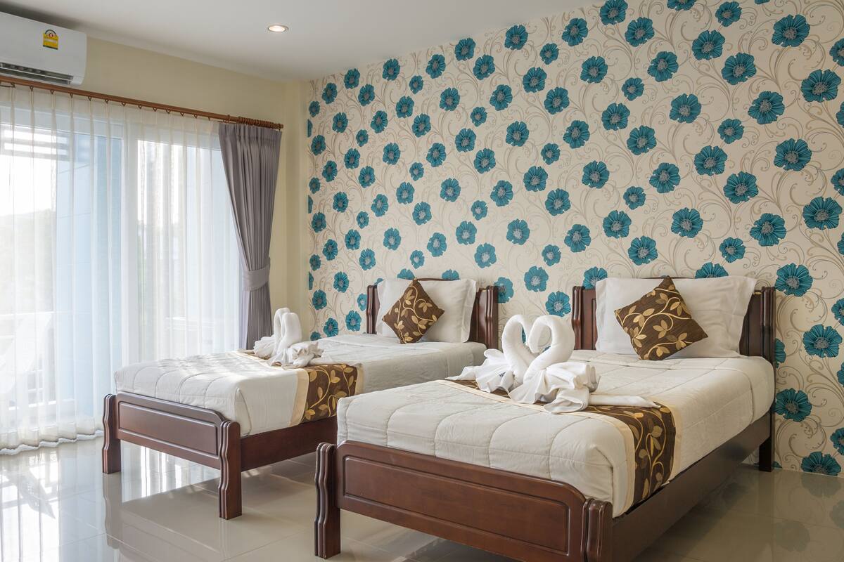 A hotel room with two beds, a large window with sheer curtains, and a floral wallpaper design.