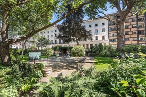 A courtyard with a central fountain, surrounded by lush greenery and a building with multiple windows in the background. - Cozy 1 Bedroom Apartment near Harrods, Knightsbridge (London)