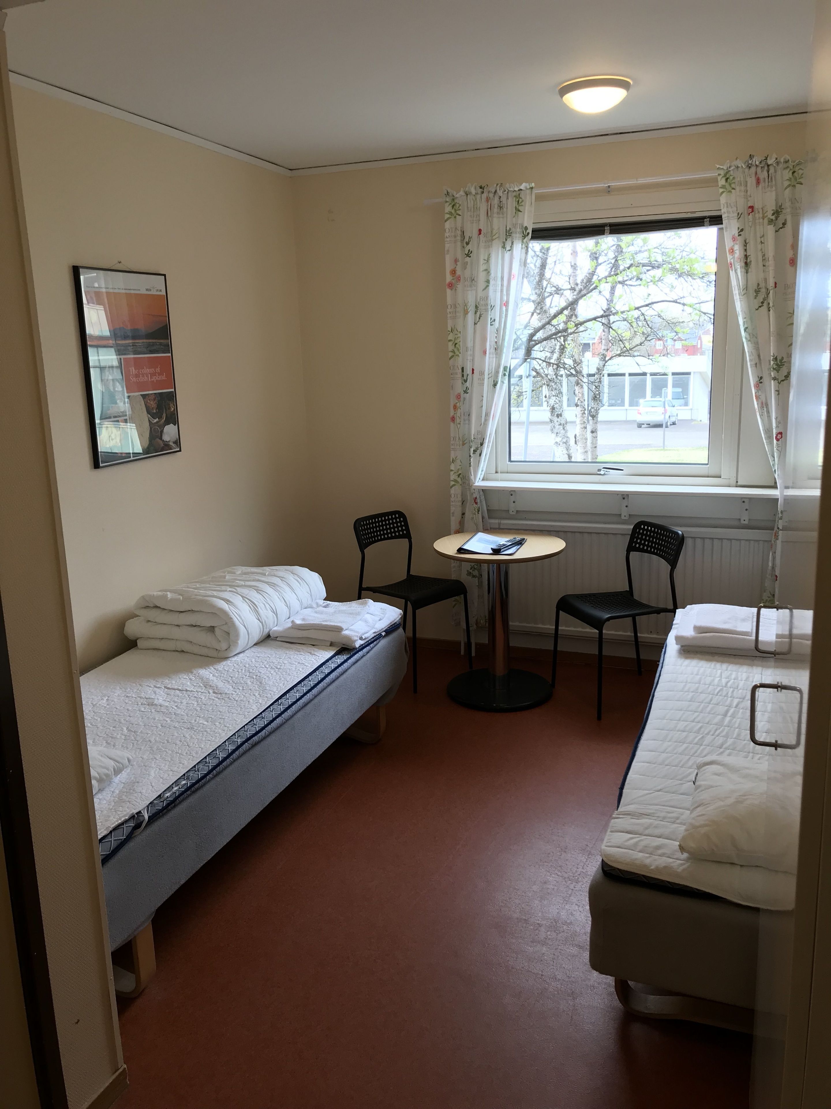A small, clean room with two single beds, a small table, two chairs, and a window with curtains.