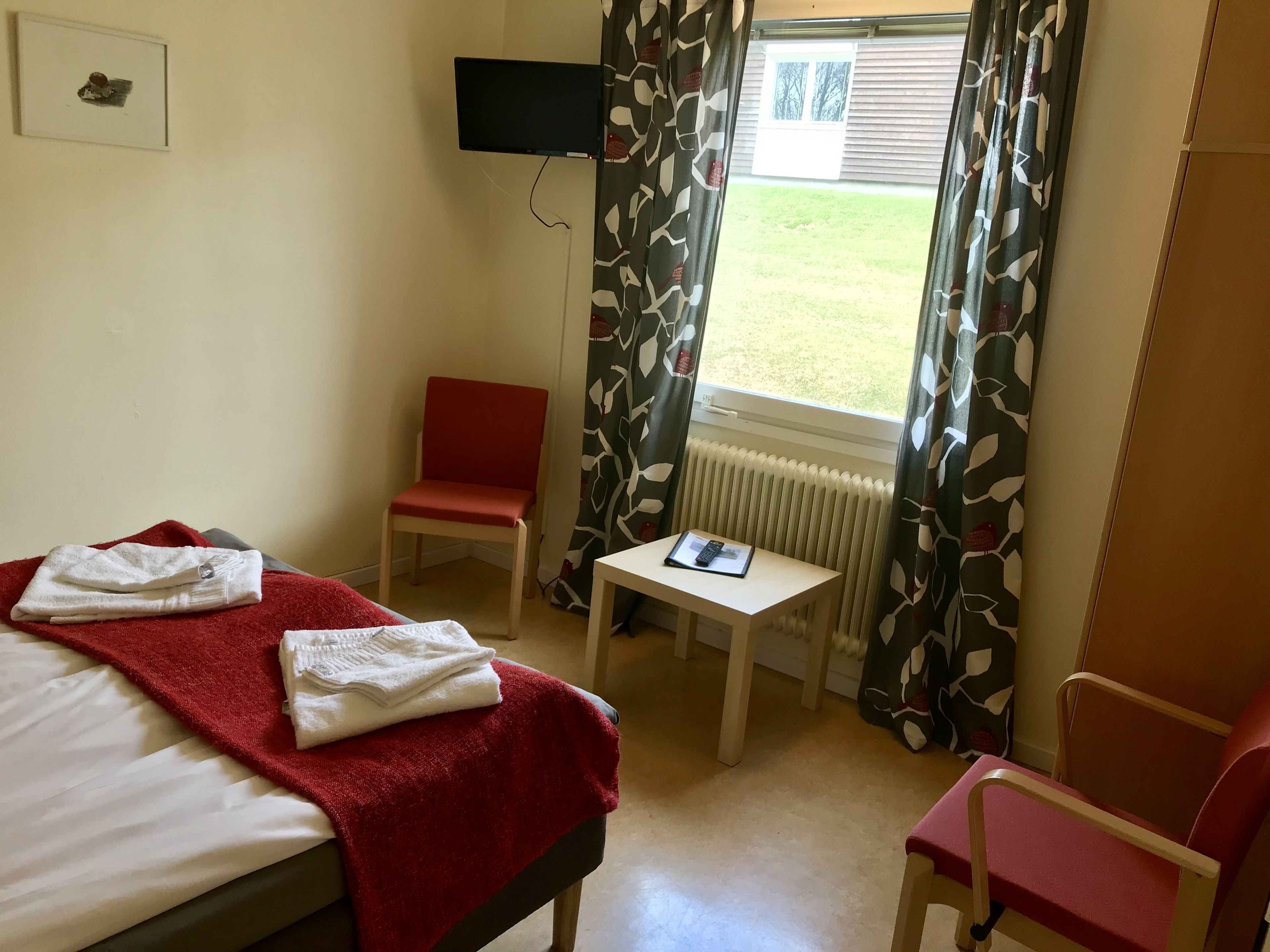 A hotel room with a bed, a red chair, a small table with a book, a TV mounted on the wall, and a window with curtains.
