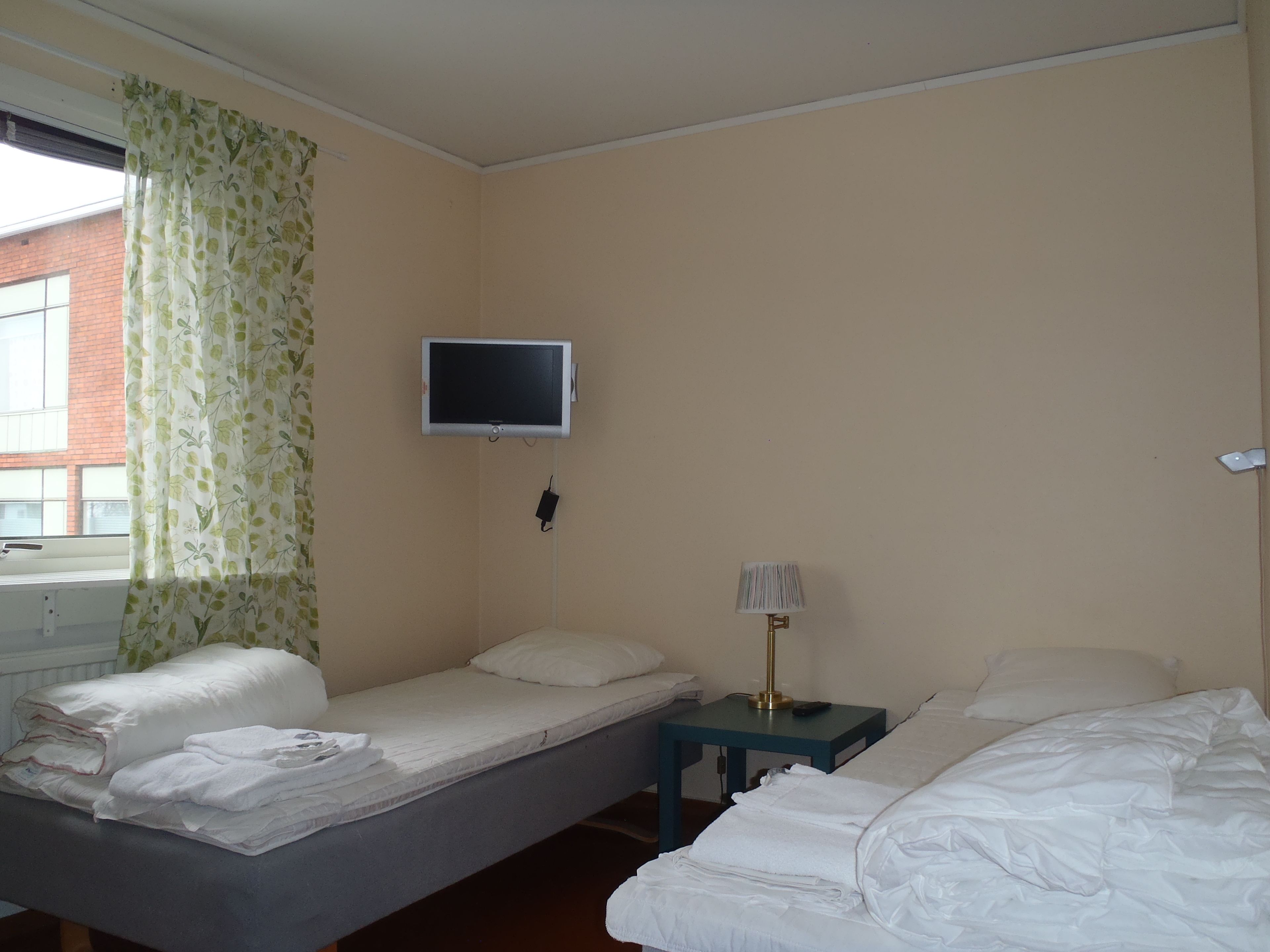 A hotel room with two single beds, a television mounted on the wall, a bedside table with a lamp, and a window with curtains.