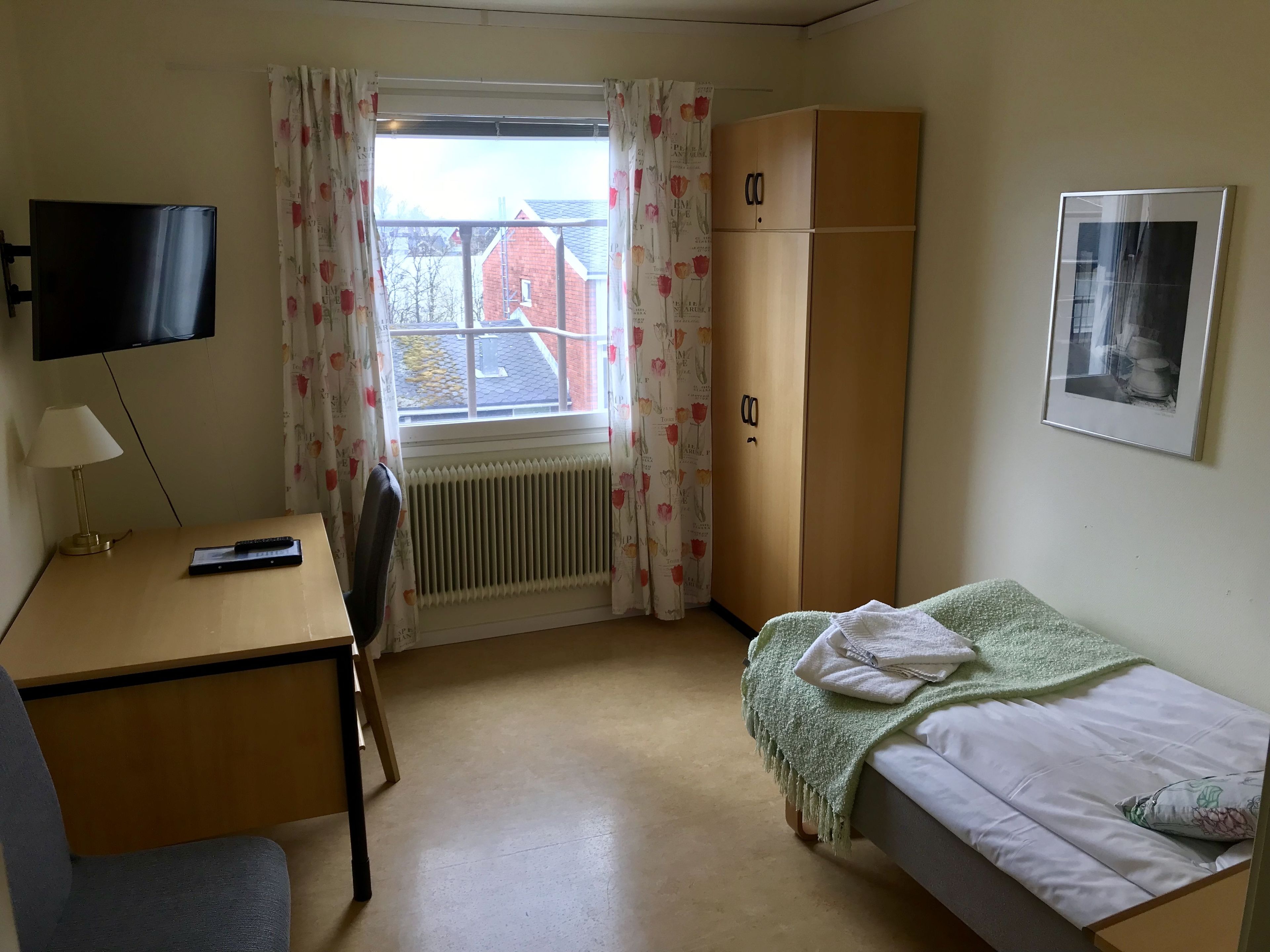 Una habitación pequeña y luminosa con una cama individual, un escritorio con silla, un radiador, un armario y un televisor montado en la pared.