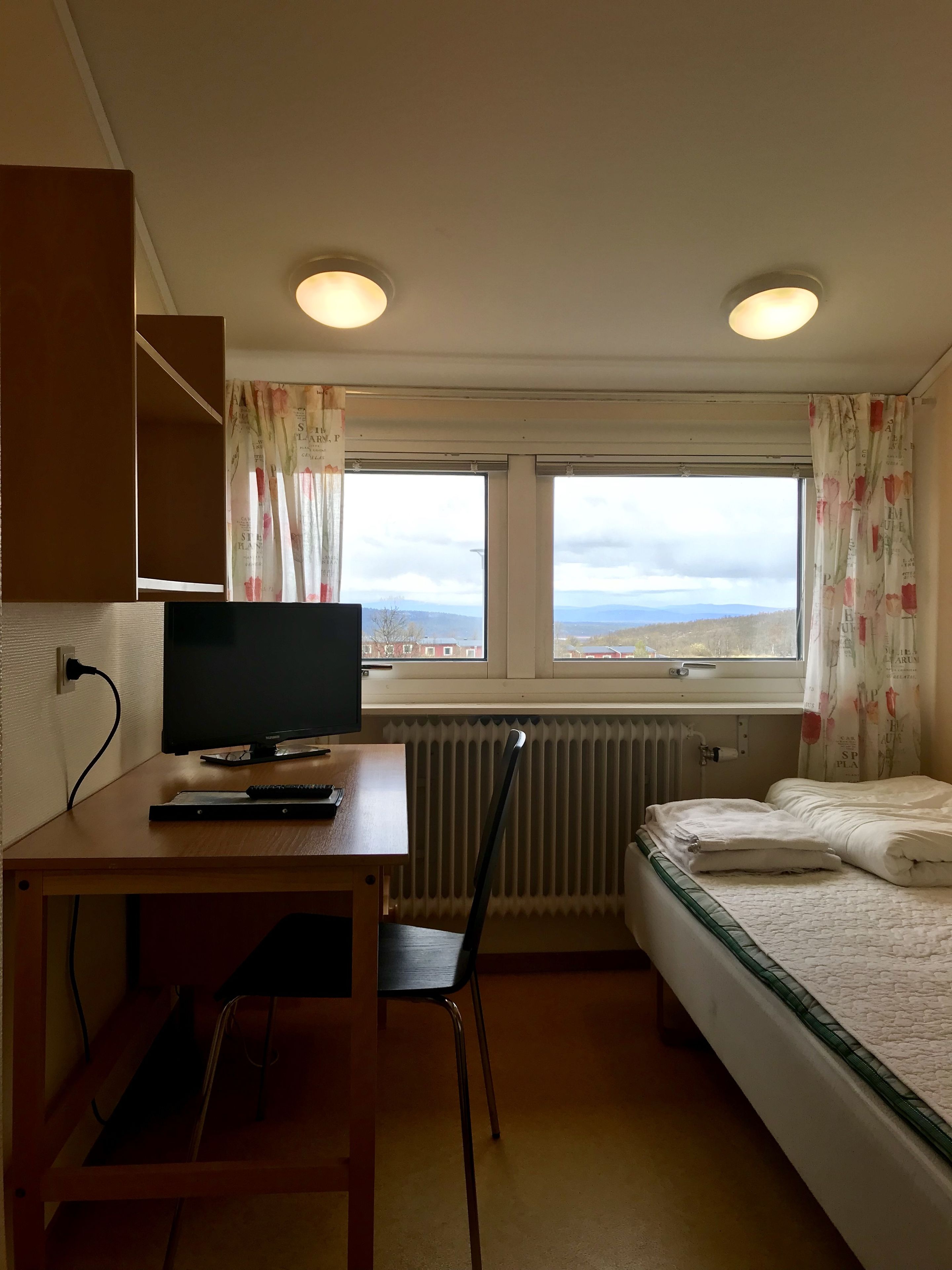 A small, well-lit room with a bed, a desk with a computer, and a chair. There are two windows with curtains, and a radiator under the windows.