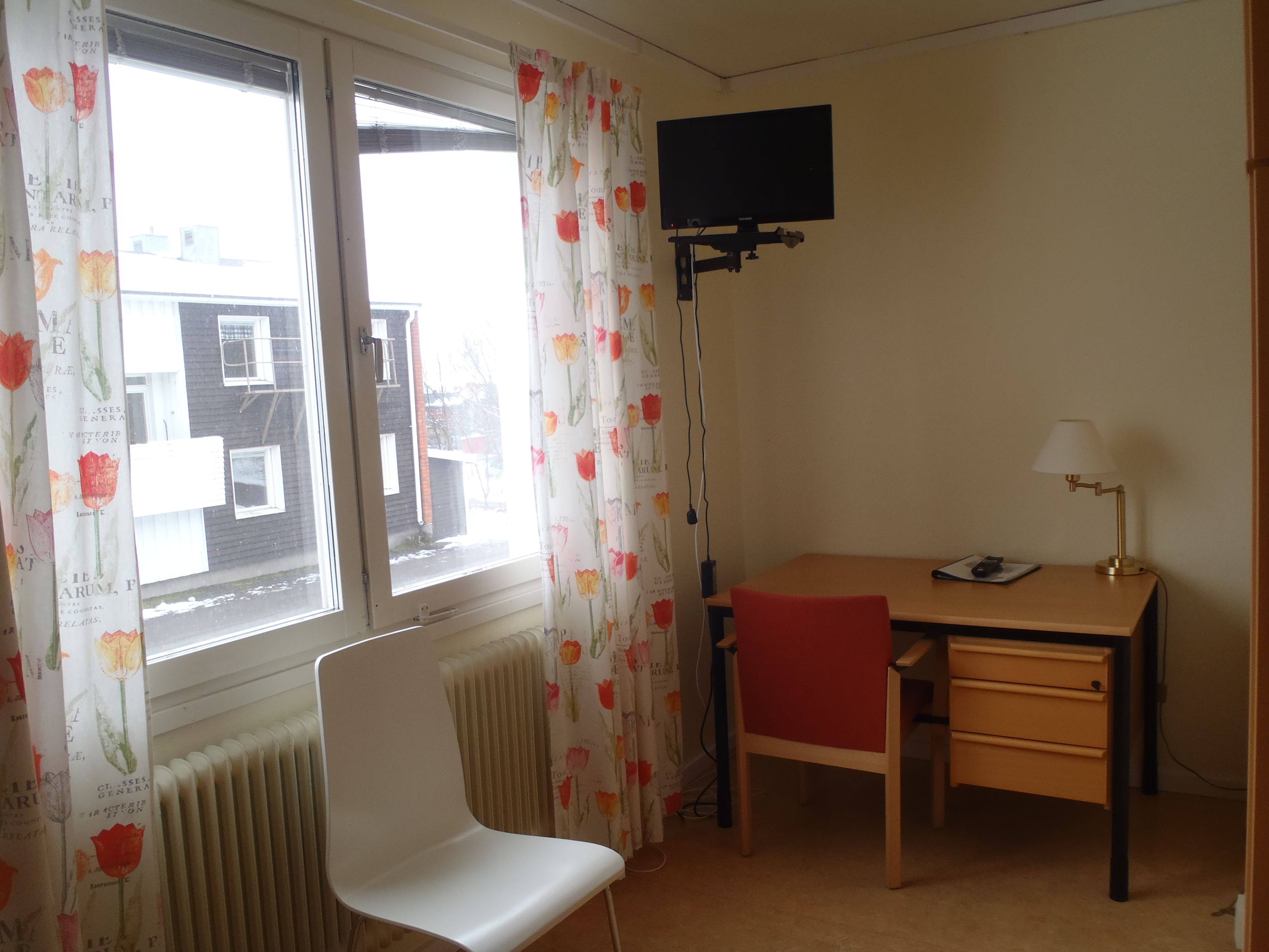 A room with a desk, chair, radiator, and a TV mounted on the wall.