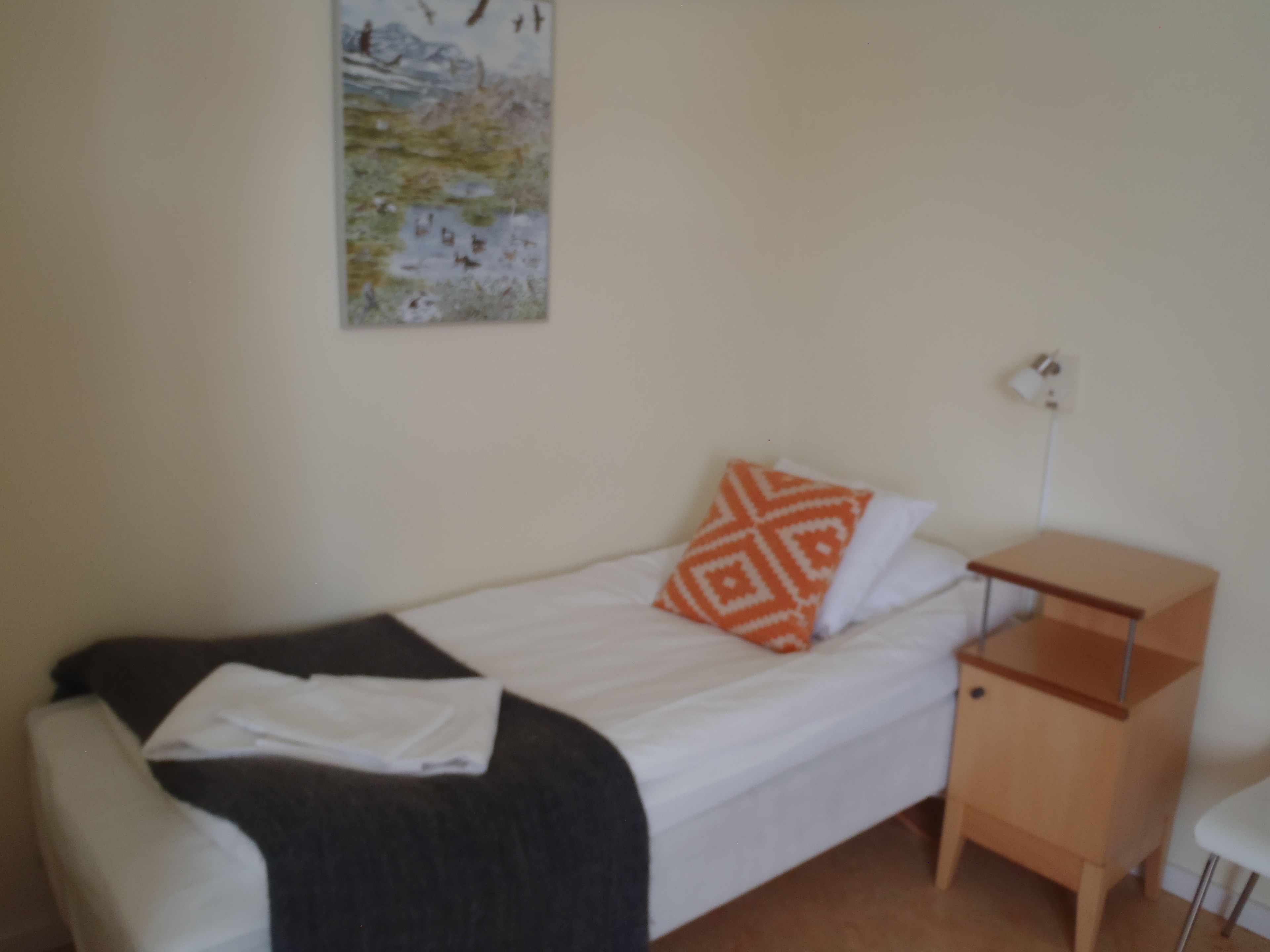 A small, neatly made bed with a white and orange patterned pillow, a wooden nightstand, and a white chair.