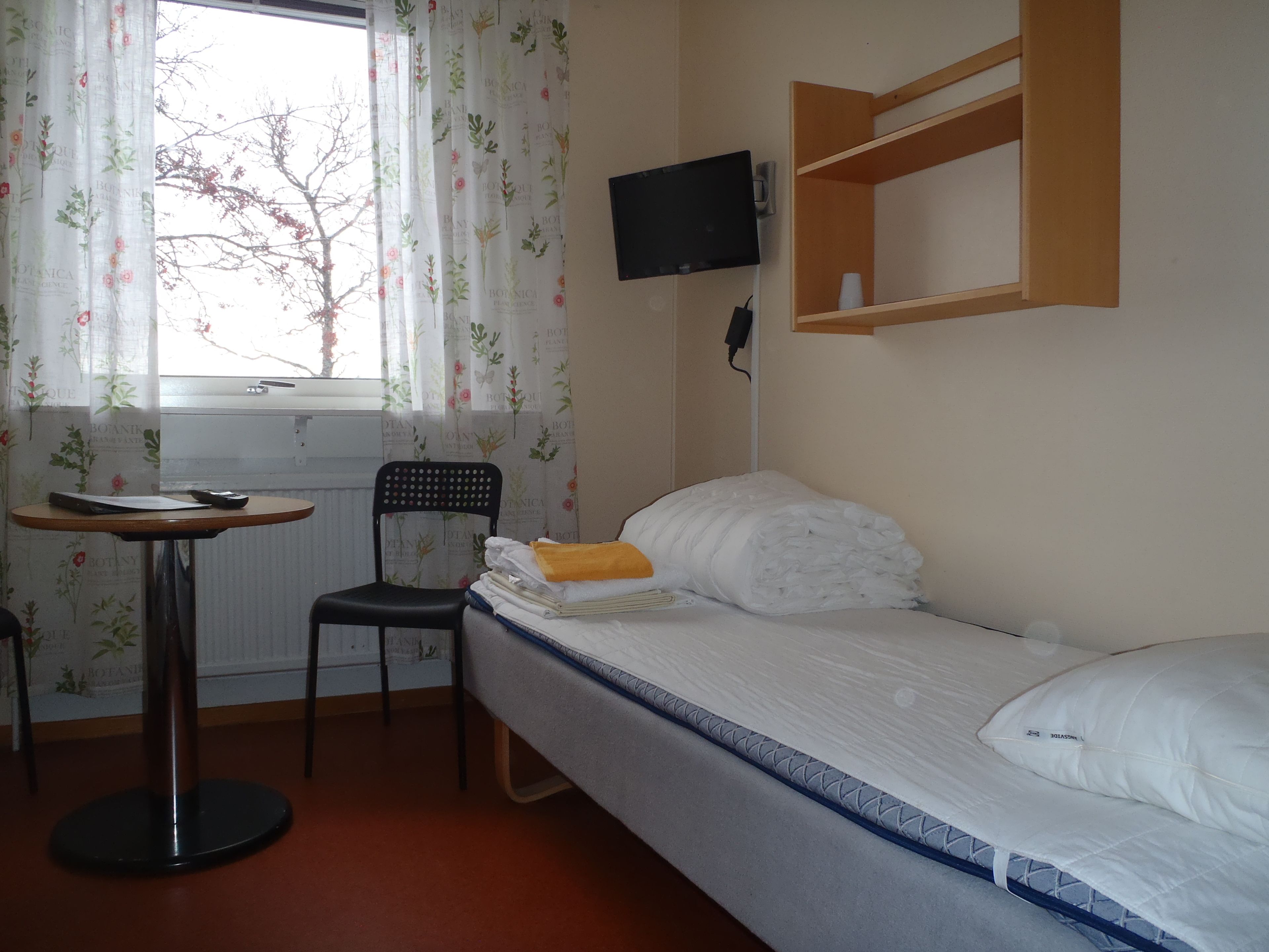 Una habitación pequeña y ordenada con una cama individual, una silla, una mesita y un televisor fijado a la pared.