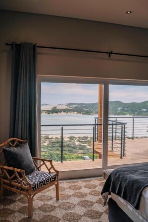 A room with a large window, a wooden chair, a bed, and a view of a balcony with a railing. - The Oyster Box Beach House (Kenton on Sea)