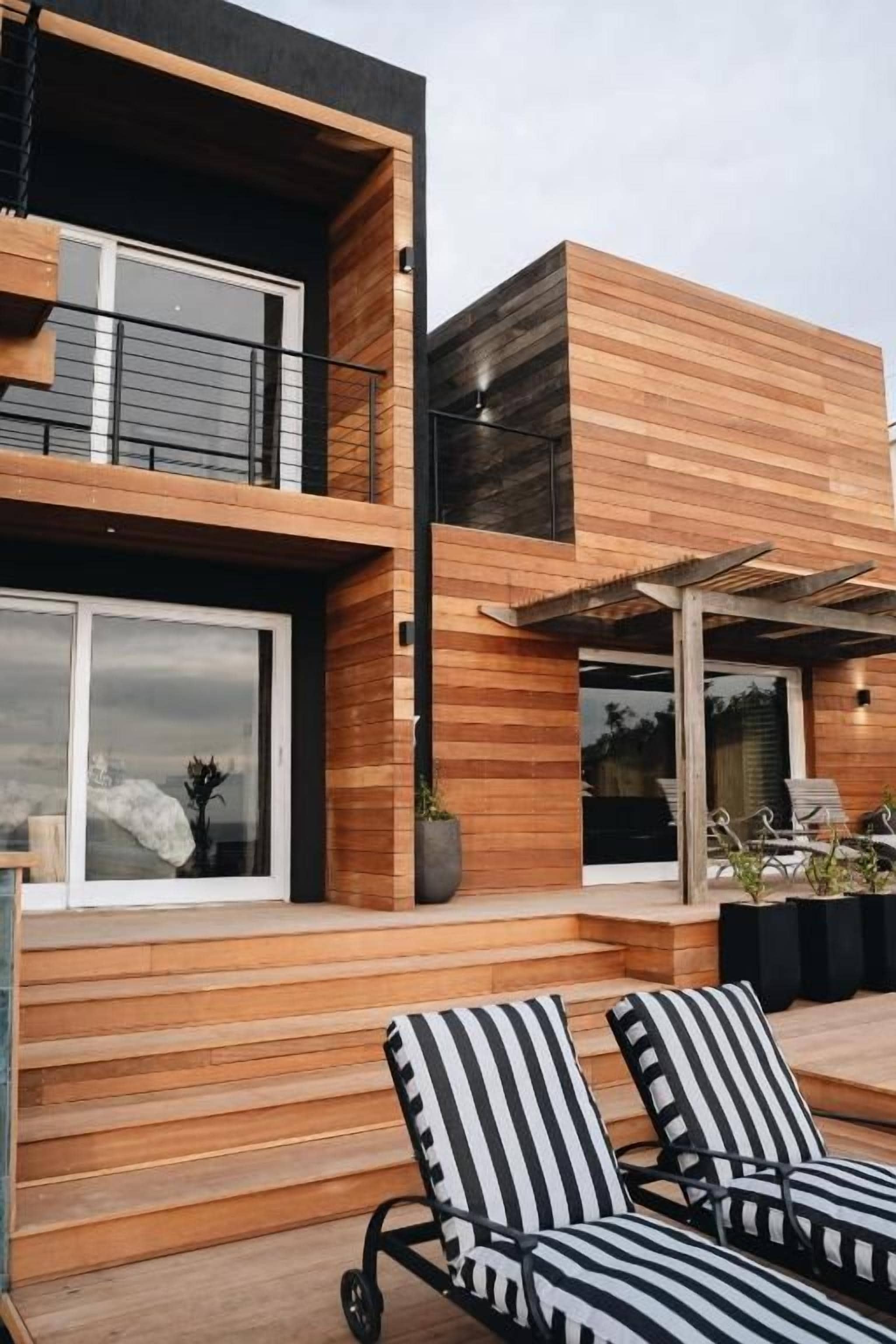 A modern two-story building with a wooden facade, a balcony, and a covered outdoor seating area with striped cushions.