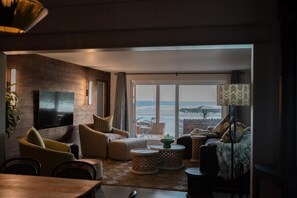 A modern living room with a large flat-screen TV, comfortable seating, a view of the beach, and a large window. - The Oyster Box Beach House (Kenton on Sea)