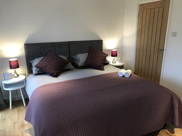 Una cama bien hecha con un edredón oscuro, dos almohadas y dos toallas enrolladas sobre la cama. Se observan dos mesitas de noche con lámparas y una puerta de madera al fondo.
