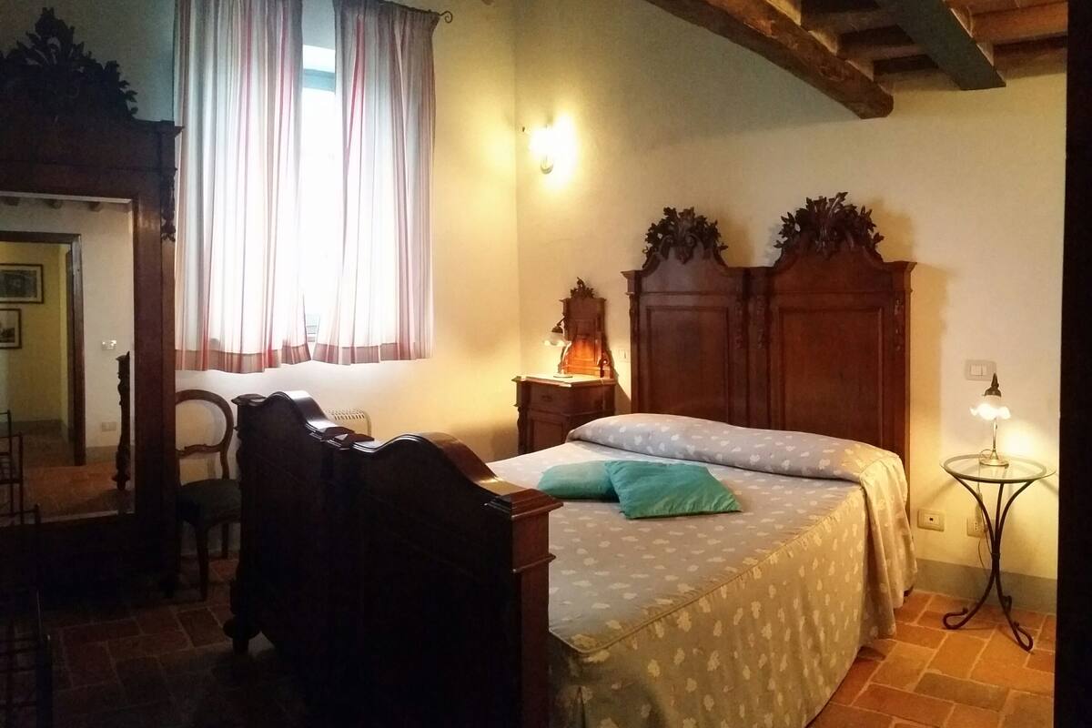A bedroom with a wooden bed, a bedside table with a lamp, a chair, and a window with curtains.