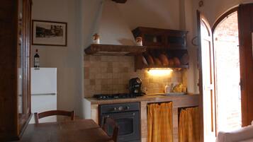 Eine Küche mit Herd, Backofen und Kühlschrank. Ein Esstisch mit Stühlen und ein Fenster mit Vorhängen sind ebenfalls vorhanden.
