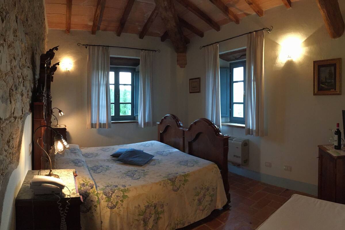 A bedroom with a bed, a bedside table with a lamp, a window with curtains, and a wall-mounted picture.