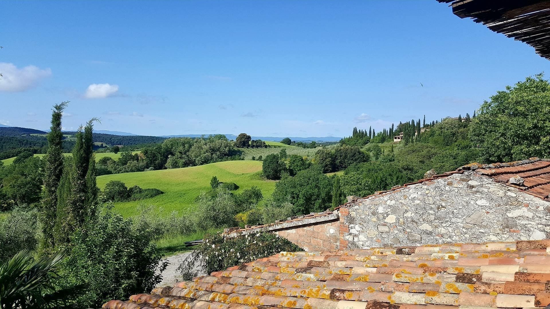 A rural landscape with rolling green fields, trees, and a stone building with a tiled roof.