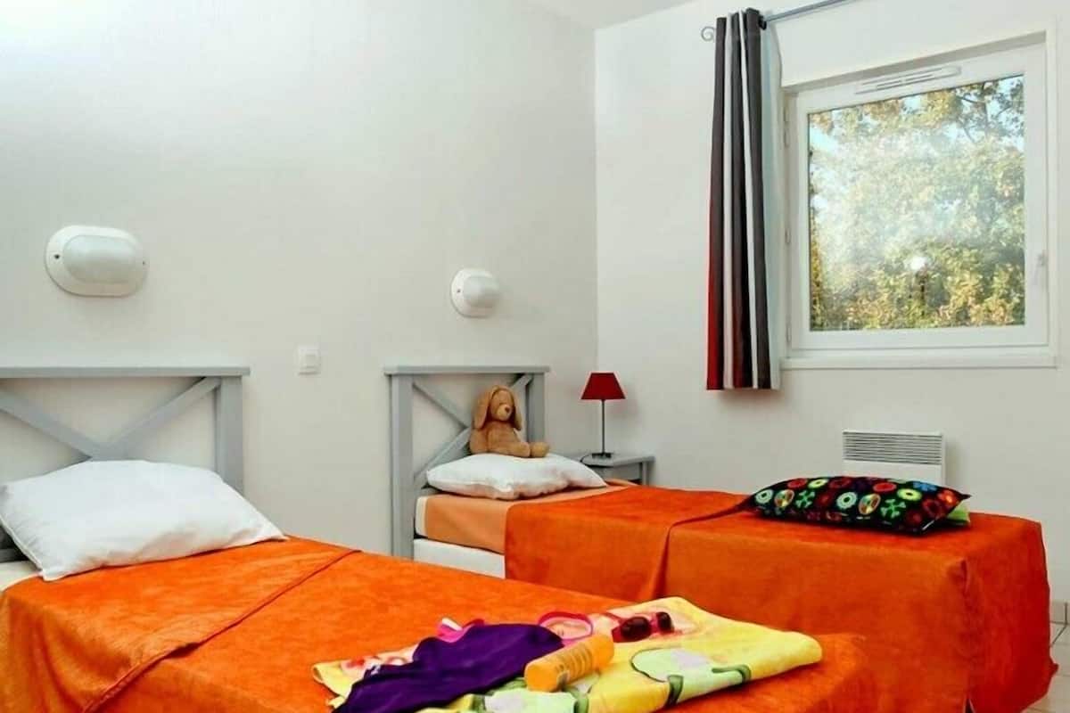 Habitació amb dos llits, cadascun amb un llençol taronja, una taula petita amb una làmpada i una finestra amb cortines.