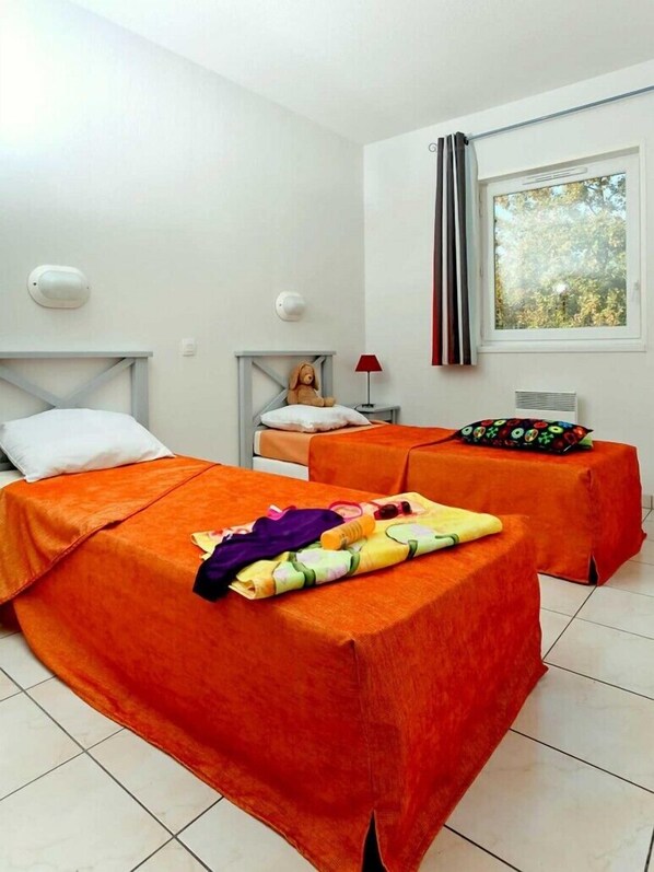 Habitación de hotel con dos camas, una ventana con cortinas, una mesita de noche con lámpara y un osito de peluche sobre una de las camas.