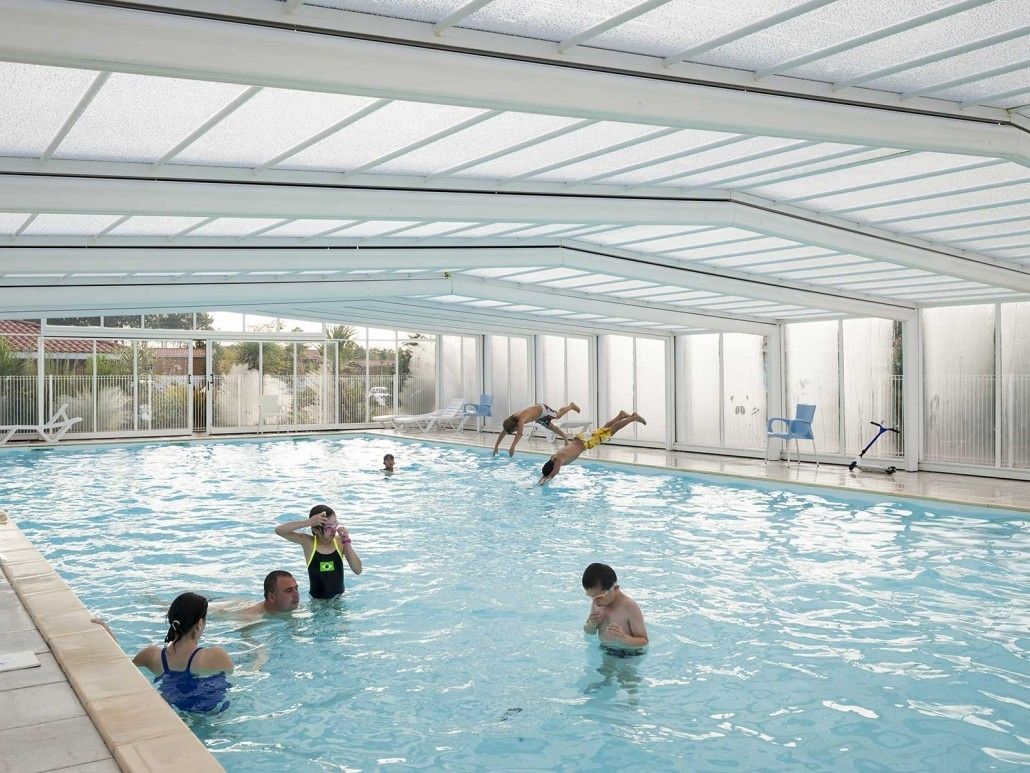 An indoor swimming pool with a glass ceiling, clear poolside chairs, and a view of the outside.