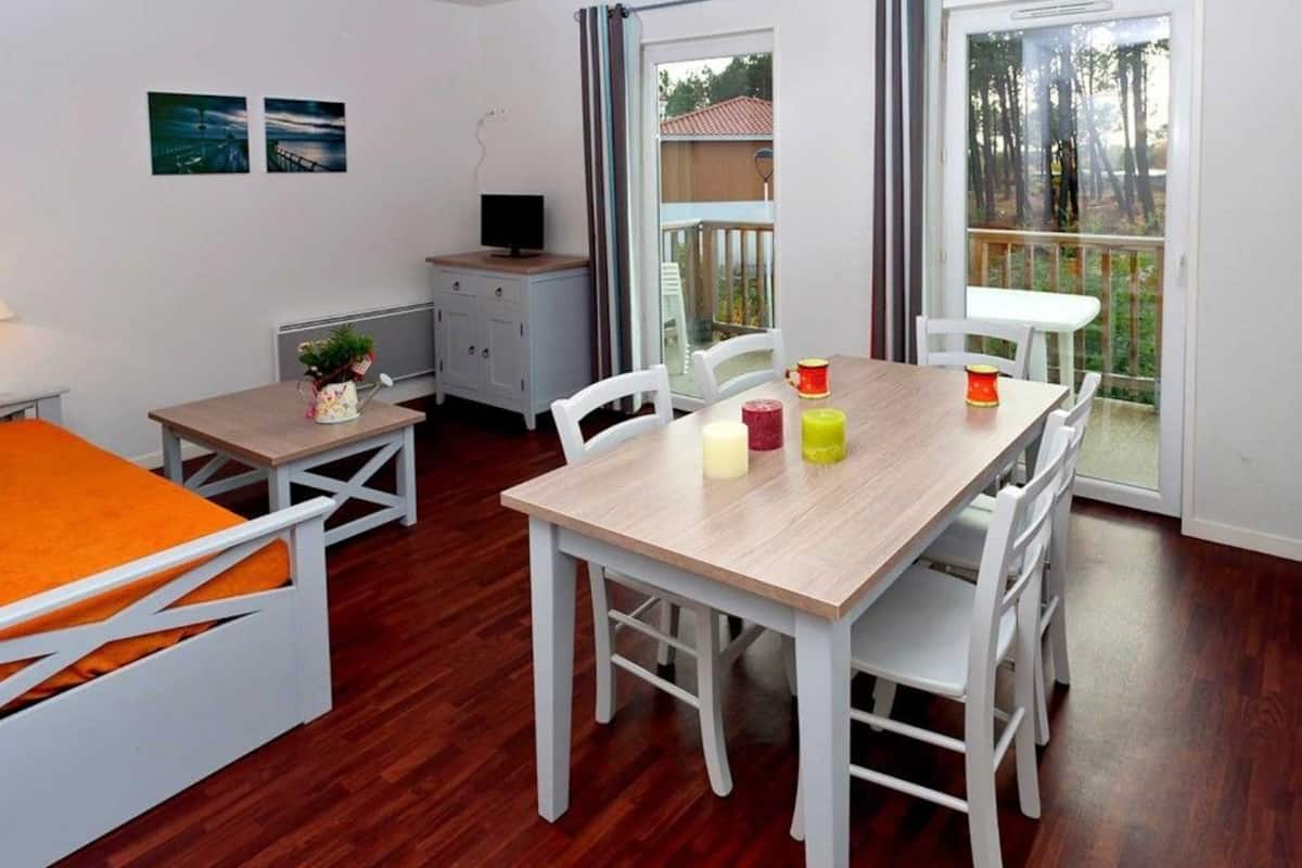 Una habitació amb taula de dinar de fusta, cadires blanques, un armari petit de color blanc i un llit amb manta taronja.