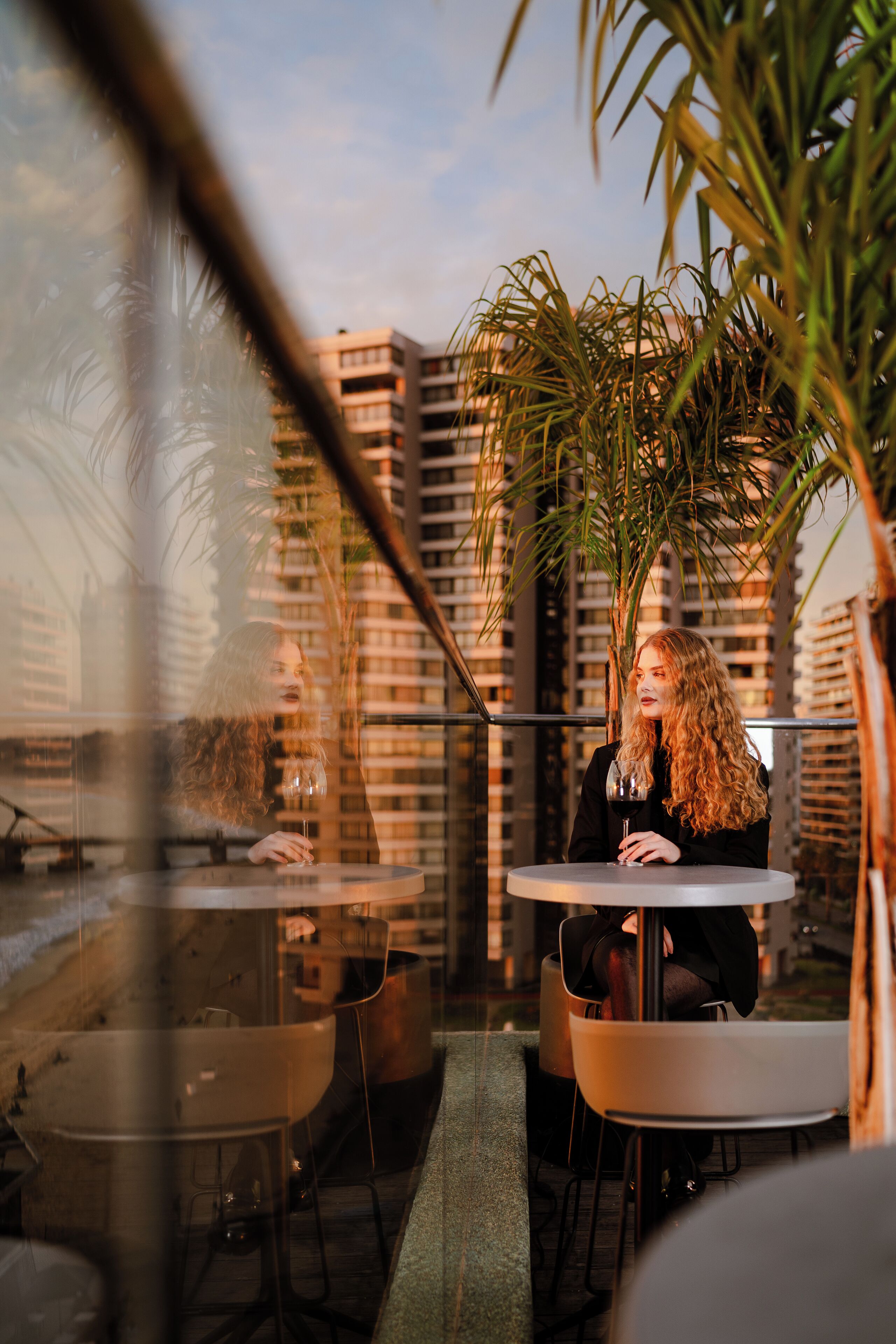 Una persona sentada en una mesa con una copa de vino, con vistas a un paisaje urbano con edificios altos y palmeras.