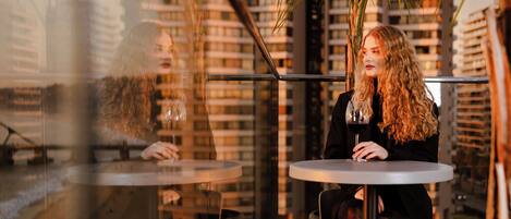 Una persona sentada en una mesa con una copa de vino, con vistas a un paisaje urbano con edificios altos y palmeras.