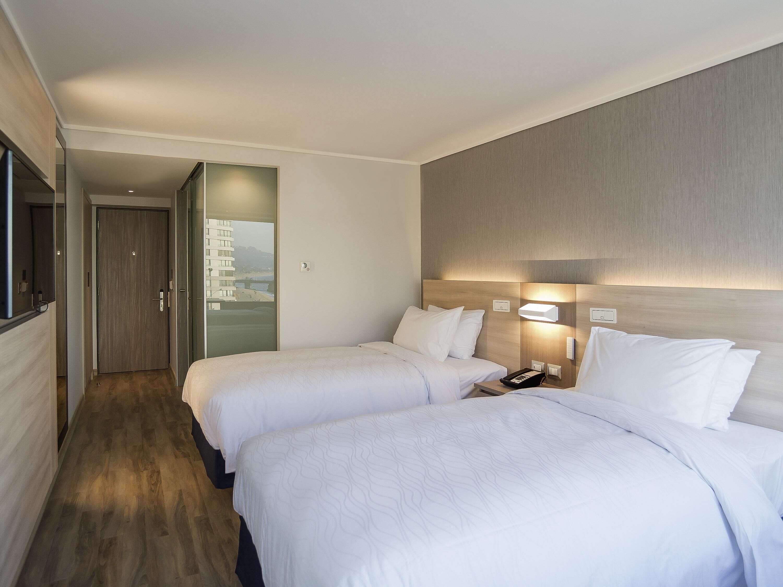 Ein Hotelzimmer mit zwei Betten, Holzboden, Blick durch eine Tür und einem Nachttisch mit Telefon.