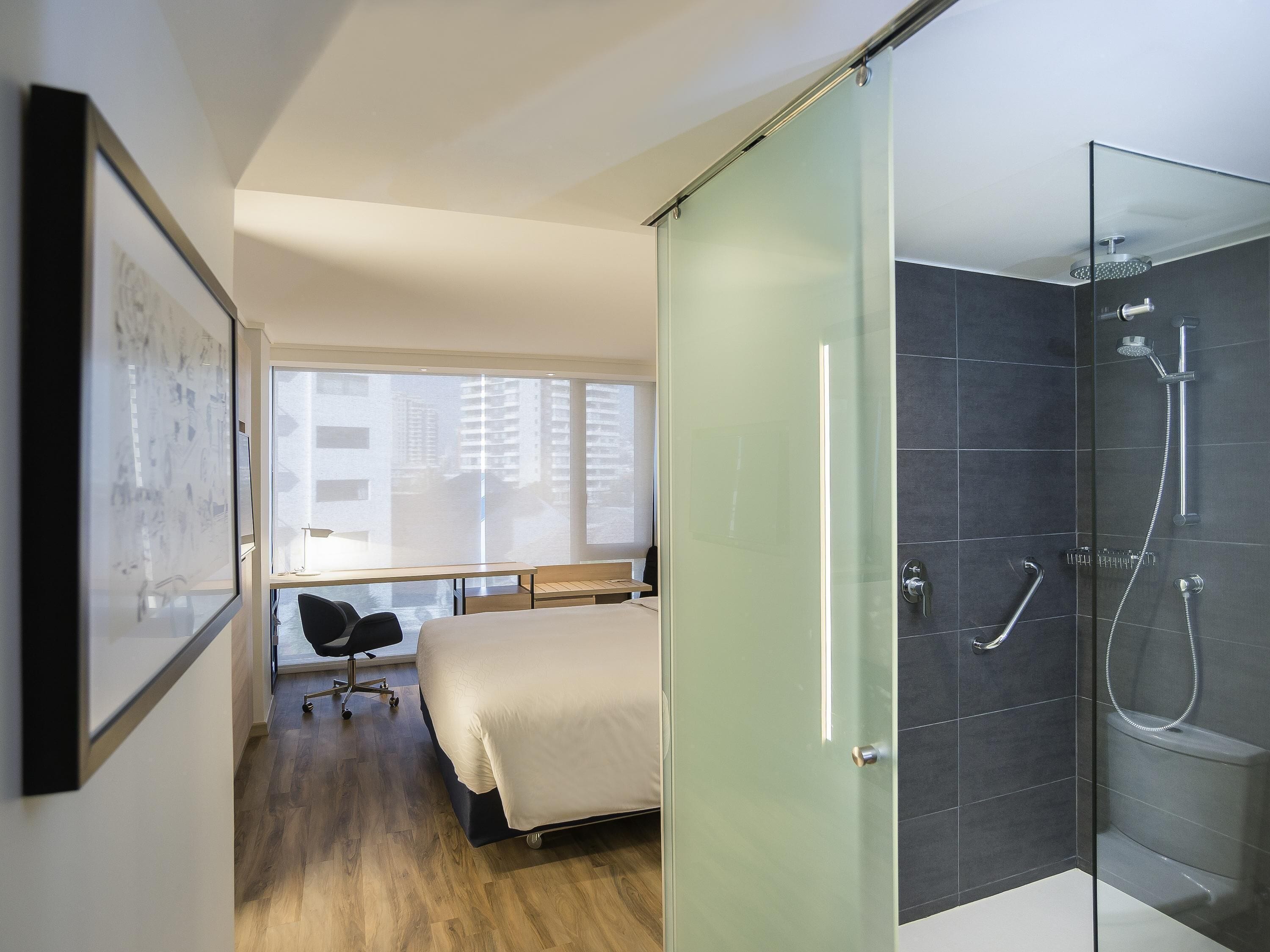 A modern hotel room with a bed, a desk, a chair, a glass shower enclosure, and a view of buildings through the window.