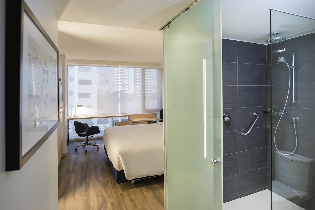 A modern hotel room with a bed, a desk, a chair, a glass shower enclosure, and a view of buildings through the window.