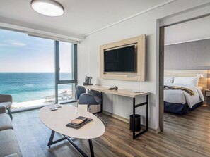 A modern hotel room with a large window offering a sea view, a desk with a chair, a bed with a headboard, and a mounted television.