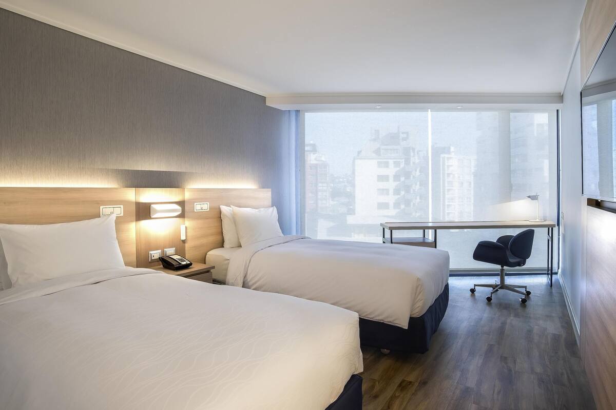 A hotel room with two beds, a desk, a chair, and a large window with a city view.