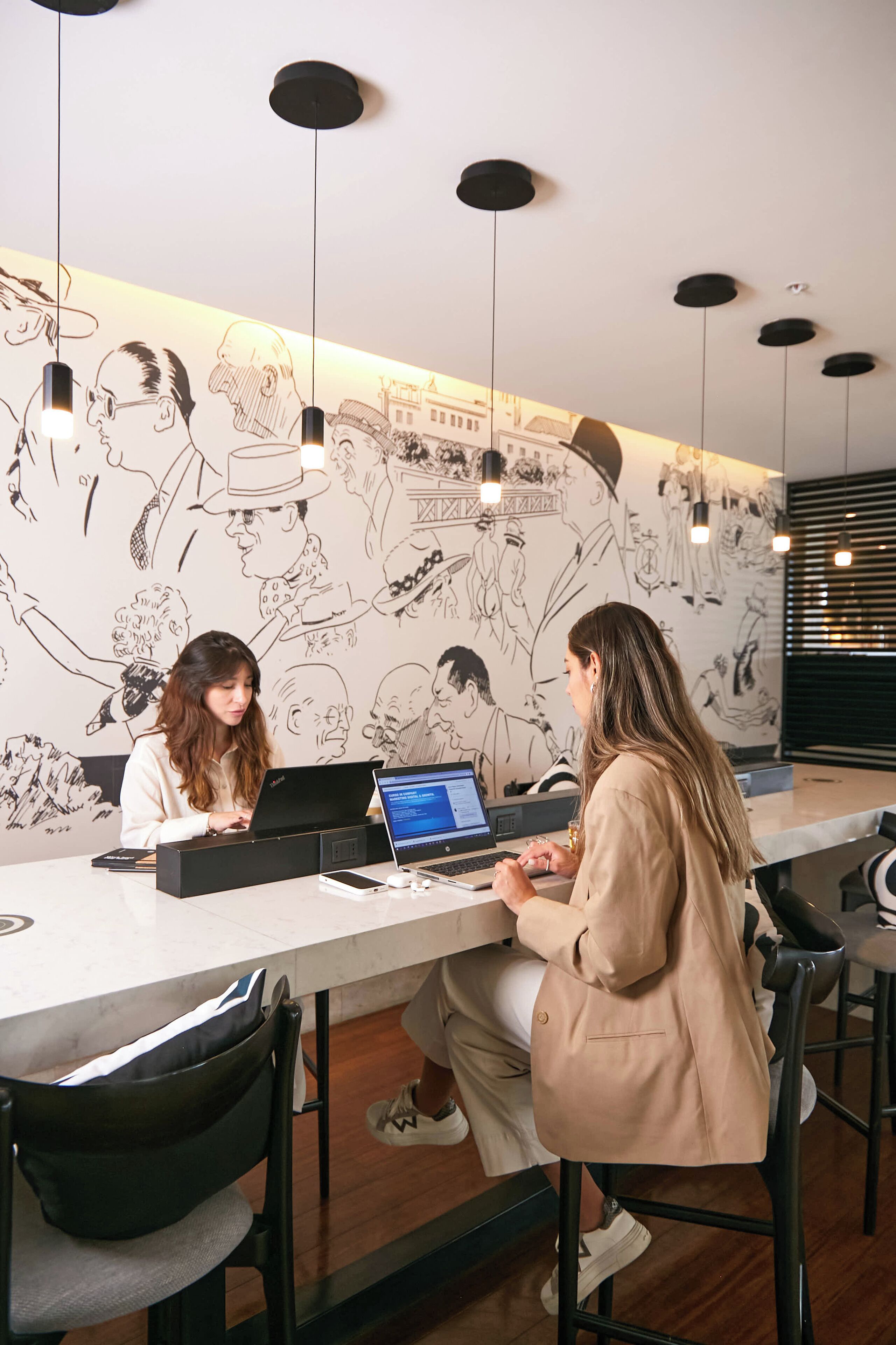 Two individuals seated at a long table with laptops, in a room with a mural of people and buildings on the wall, and pendant lighting.