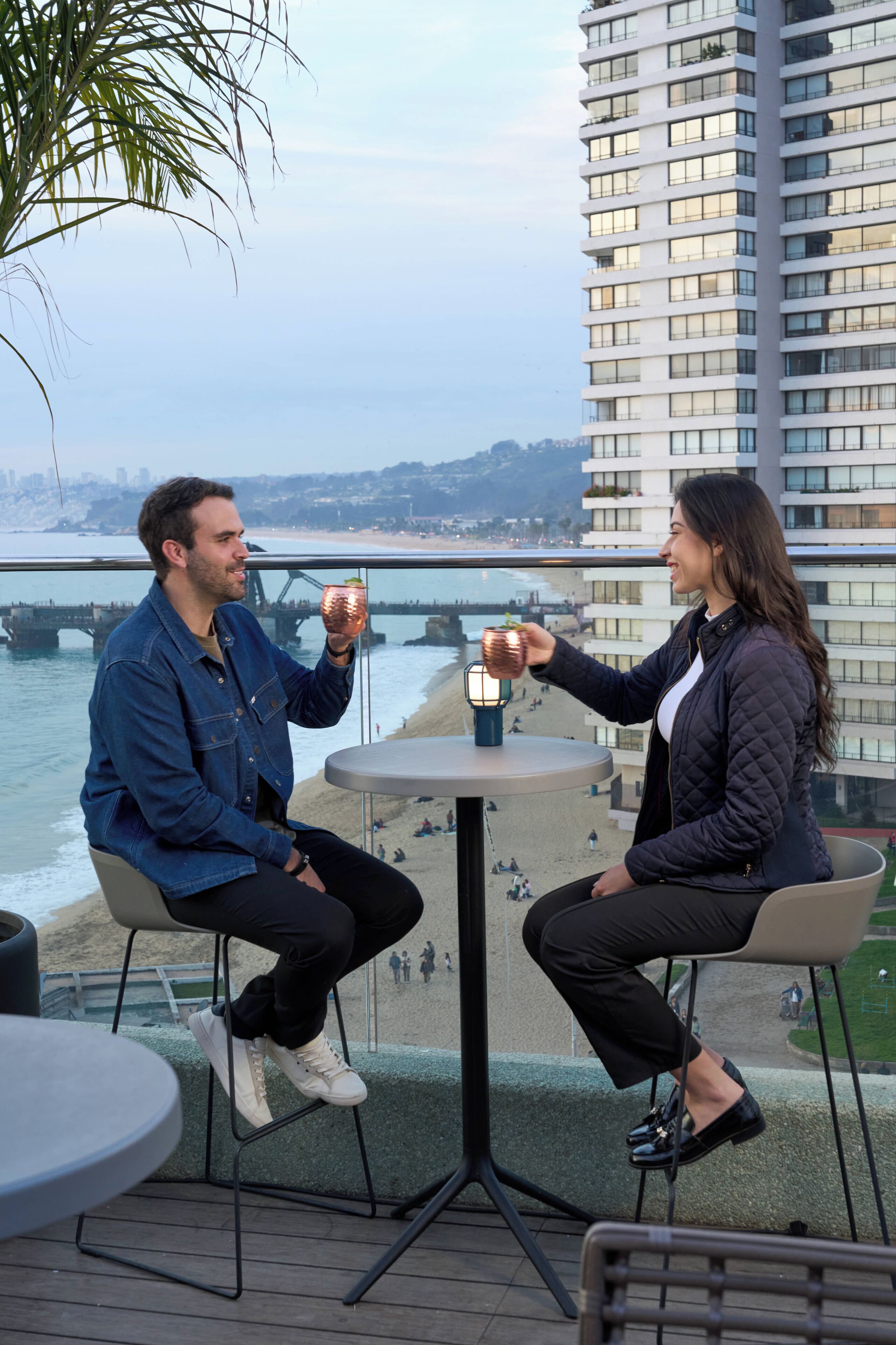 Deux personnes sont assises à une table, avec vue sur la plage et un pont en arrière-plan.
