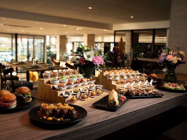 A buffet with various dishes, including burgers, salads, and desserts, arranged on a wooden table in a well-lit room with large windows. - Pullman Viña del Mar San Martín (Vina del Mar)