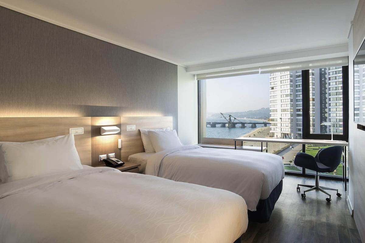 A hotel room with two beds, a nightstand, a desk, and a large window with a view of a bridge and buildings.