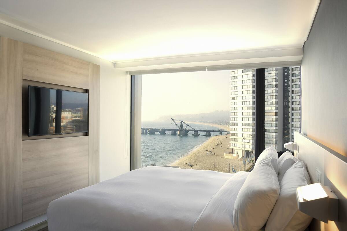 A modern hotel room with a large bed, a television, and a view of a bridge and beach.