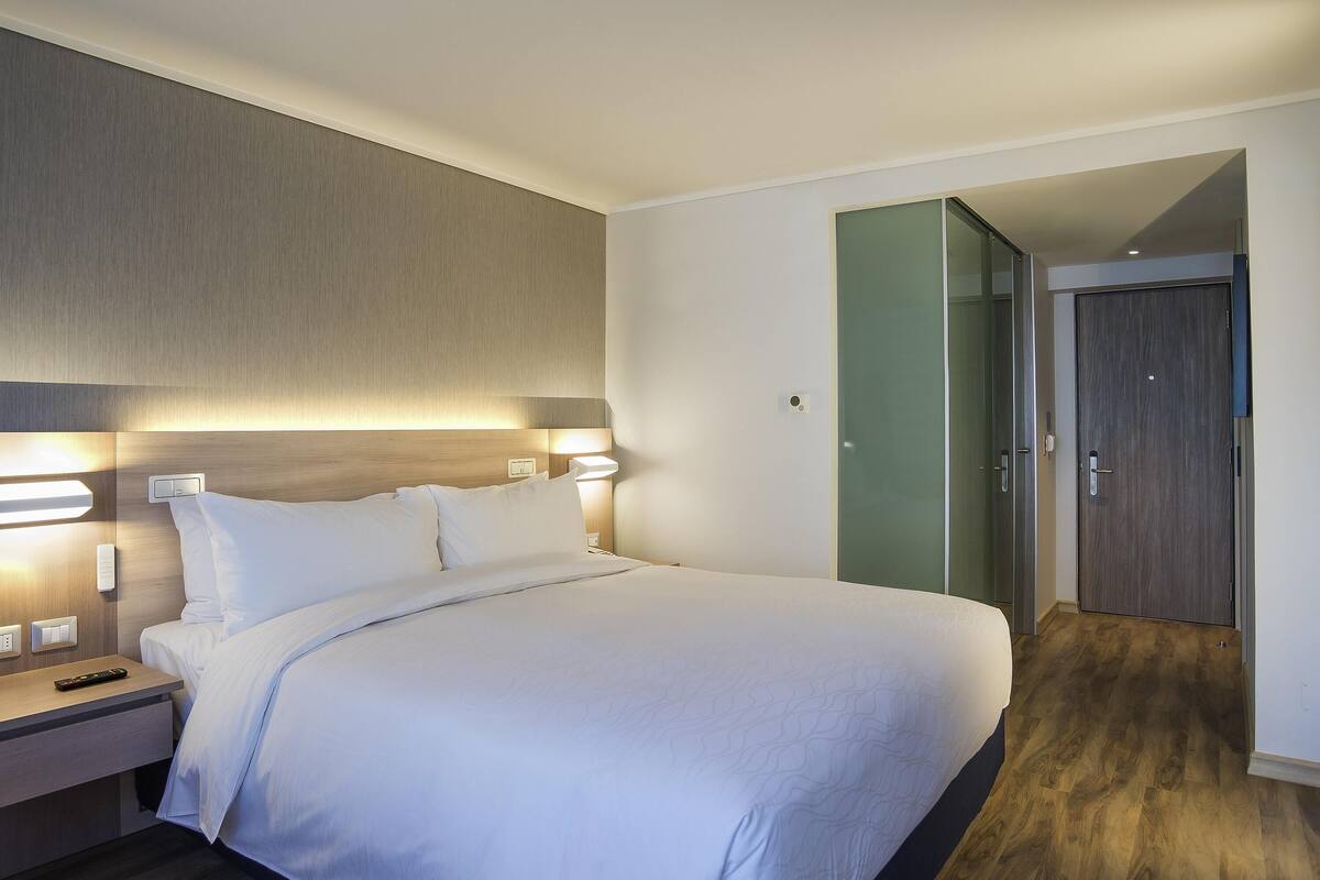 A hotel room with a bed, bedside lamps, a nightstand, a wooden floor, and a door.