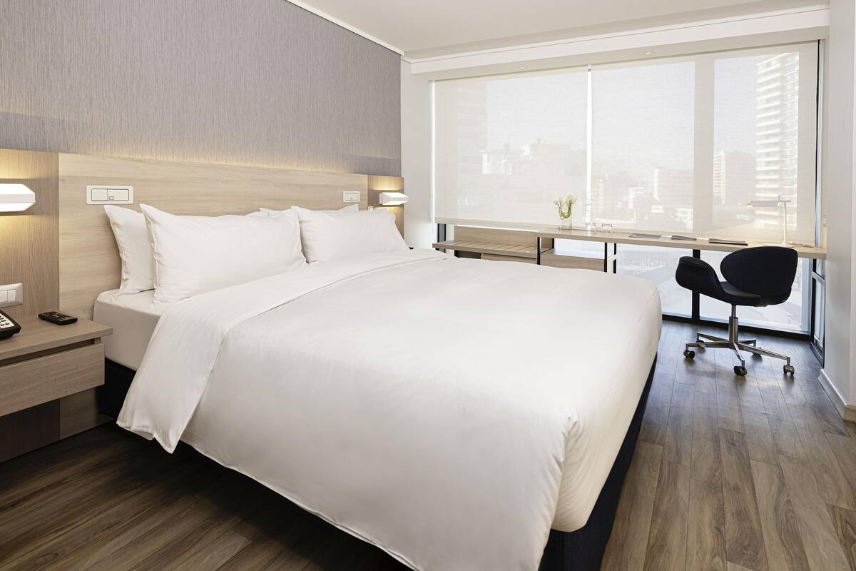 A modern hotel room with a large bed, bedside tables, a desk with a chair, and a large window offering a city view.